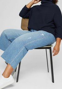 Jeans a gamba larga di colore azzurro chiaro con dettagli strappati, orlo sfilacciato, abbinati a un dolcevita blu navy scuro e sneakers bianche, mentre è seduto su una sedia.