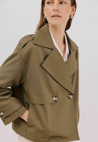 Oliwkowa kurtka o podwójnym zapięciu, z dużym kołnierzem i błyszczącymi guzikami. Materiał wydaje się gładki i ma uporządkowany krój.