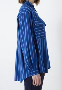 Donna che indossa una camicia blu svasata con righe verticali azzurre, polsini abbottonati e tasche sul petto, mostrata di lato.