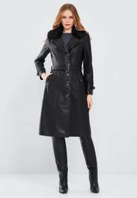 Cappotto in pelle nera con collo in pelliccia sintetica, vita cinturata, chiusura con bottoni, tasche e design fino al ginocchio. Texture liscia e forma aderente.