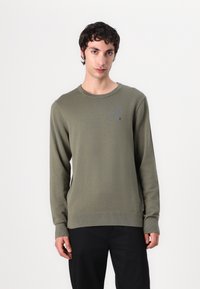 Pulover tricotat verde măsliniu, cu mâneci lungi și manșete cu nervuri. Prezintă un logo mic pe piept. Stilizat cu pantaloni negri.