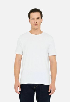 Junger Mann mit kurzem dunklem Haar, der ein einfaches weißes T-Shirt mit kurzen Ärmeln und dunklen Jeans trägt, steht gegen einen weißen Hintergrund.