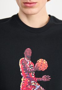 Detailní záběr osoby v černém shirtu s barevným pixelovaným grafickým motivem basketbalisty držícího basketbal.