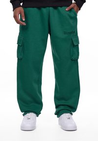 Grønne cargo sweatpants med sidelommer, lavet af blødt stof. Har elastisk talje og justerbare manchetter. Kombineret med hvide sneakers.