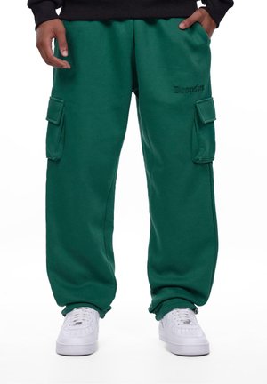 Pantalons de survêtement cargo verts avec poches latérales, fabriqués en tissu doux. Comprend une taille élastique et des poignets ajustables. Associés à des baskets blanches.