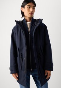 Navy blue parka jacket with a hood, featuring zip and button closures, drawstring waist, and two front pockets. Worn over a navy zip-up sweater.