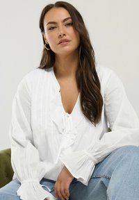 White blouse with a V-neck and ruffled detailing, featuring long, voluminous sleeves with elastic cuffs. Paired with blue jeans.