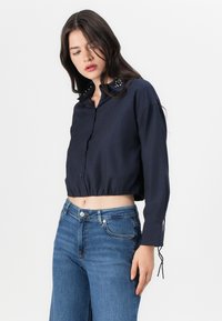 Camisa cropped azul-marinho com fecho de botão, apresentando detalhes de colarinho decorativo e mangas longas com cordão nos punhos. Usada com jeans azuis.