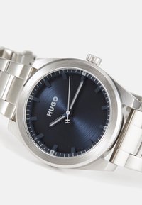 Reloj de acero inoxidable con una esfera redonda de color azul marino, marcadores de hora y agujas plateadas. Cuenta con una pulsera de eslabones cepillados y una corona en el lateral.