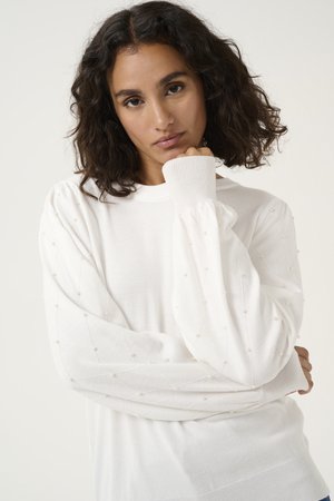 Femme aux cheveux bouclés mi-longs portant un pull blanc à manches longues avec des détails en perles sur les manches, reposant son menton sur sa main.