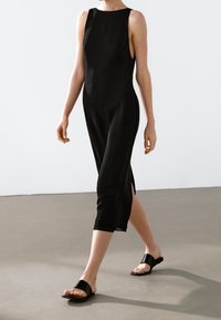 Robe noire sans manches avec fentes latérales, fabriquée en tissu lisse. Associée à des sandales noires à enfiler présentant une large bride et un design minimaliste.