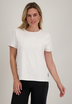 Vrouw met blond haar die gouden creooloorbellen draagt, een effen wit kort mouw shirt en zwarte broek tegen een lichtgrijze achtergrond.