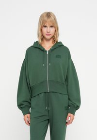 Zelená skrátená mikina s kapucňou vyrobená z m�äkkej látky, s predným zipsom, šnúrkou na kapucni, oversized rukávmi a malou akcentovou nášivkou.
