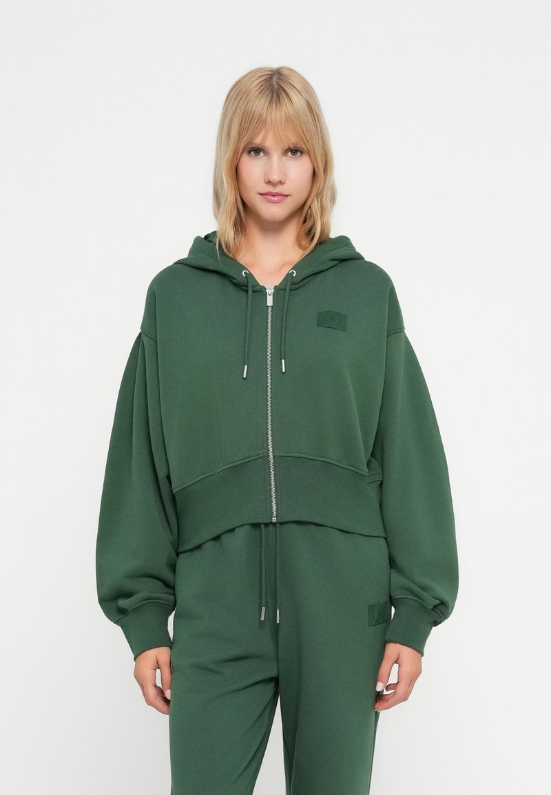 Zelená skrátená mikina s kapucňou vyrobená z mäkkej látky, s predným zipsom, šnúrkou na kapucni, oversized rukávmi a malou akcentovou nášivkou.