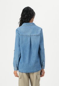 Personne aux longs cheveux foncés portant une chemise en denim bleue à manches longues et un pantalon cargo beige, vue de dos sur fond blanc.