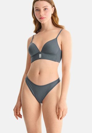 Jeune femme à la peau claire et aux longs cheveux portant un ensemble soutien-gorge côtelé gris assorti et culotte, sur un fond blanc uni.