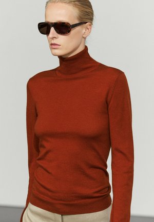 Frau mit blondem Haar, die Schildpatt-Sonnenbrillen, einen rostfarbenen Rollkragenpullover und beige Hose vor einem einfarbigen Hintergrund trägt.