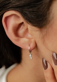 Pendiente de aro plateado con una piedra rectangular clara colgando de él, exhibido en un primer plano de una oreja y la textura de la piel.