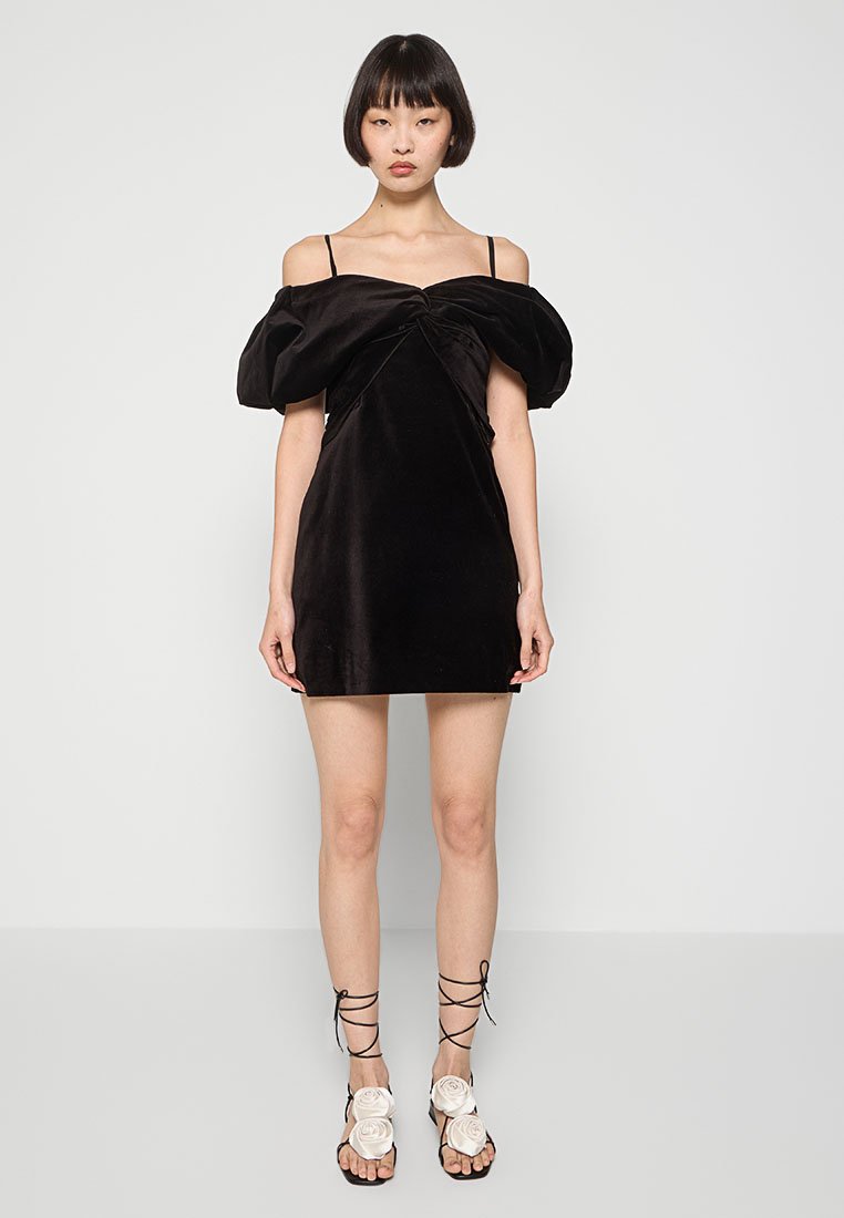 Robe mini en velours noir avec des manches bouffantes dénudées des épaules et un design de corsage torsadé. Le modèle porte des sandales à lacets avec des accents floraux.