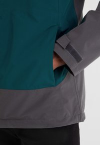 Person wearing a teal and gray jacket with hand in side pocket, showing a gray cuff with Velcro fastener.