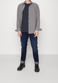 Camisa de cuadros en blanco y negro sobre una camiseta oscura, combinada con vaqueros azul oscuro y botas negras. Atuendo casual con un look en capas.
