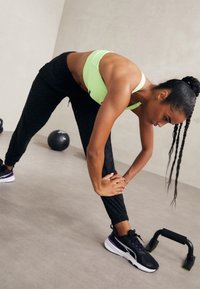 Soutien-gorge de sport vert clair et joggings texturés noirs. Une personne se penche en avant, tenant une jambe. Chaussures de sport noires et violettes, matériel d'exercice à proximité.