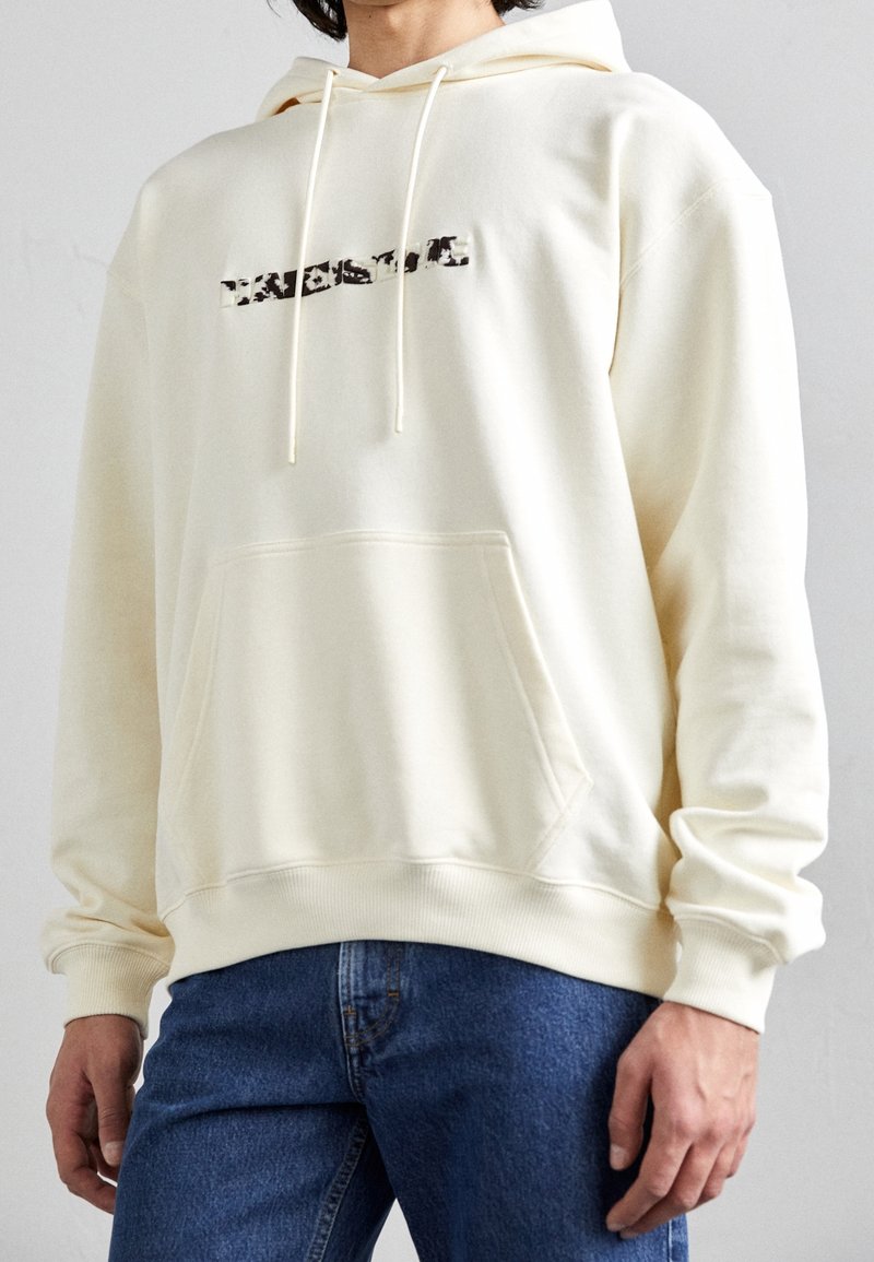 Personne portant un sweat à capuche écru avec une poche kangourou à l'avant et un texte noir déformé sur la poitrine, assorti à un jean bleu.