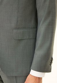 Zbliżenie na rękaw szarej marynarki i kieszeń z widocznym mankietem białej koszuli na ramieniu osoby.