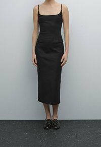 Kvinna i en svart ärmlös topp och matchande lång kjol, stående mot en enkel ljusgrå bakgrund med svarta skor.