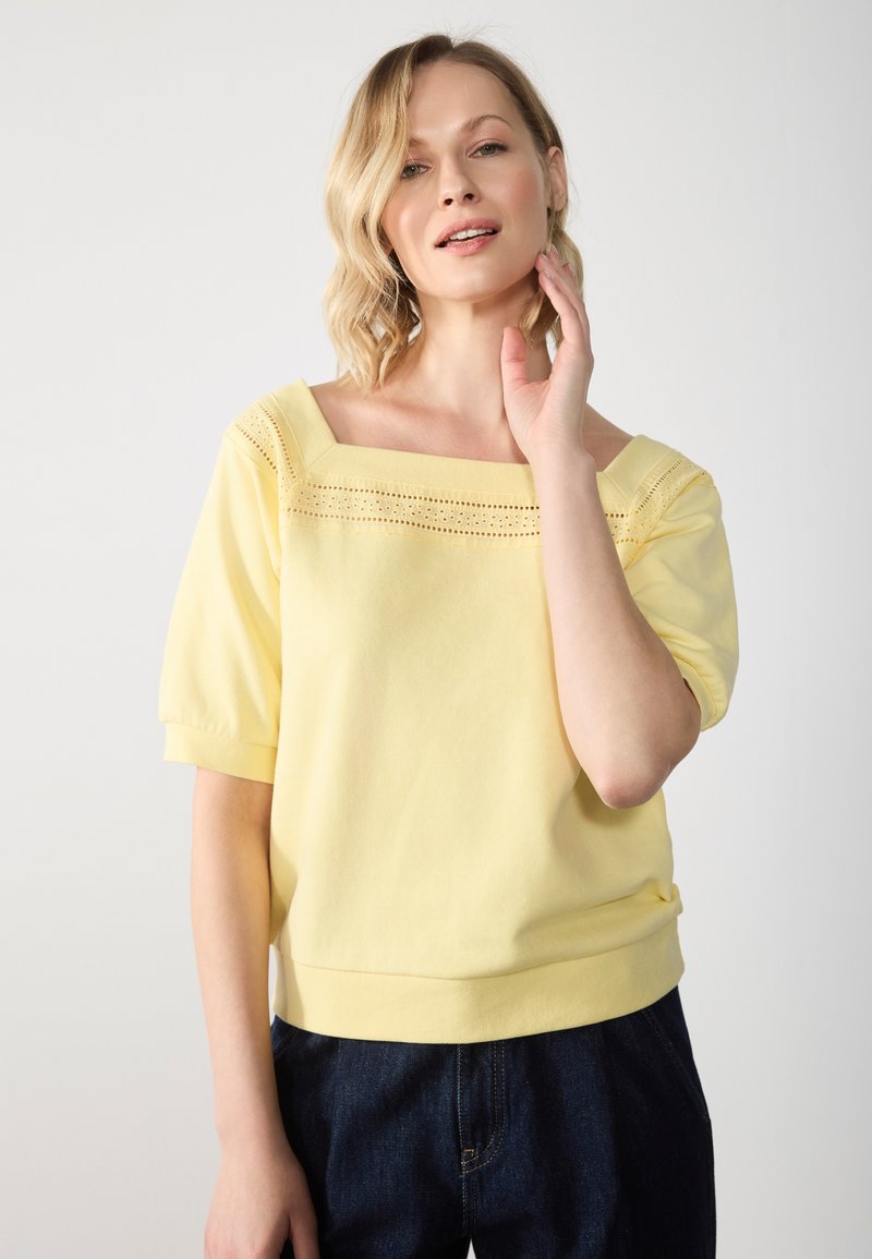 Top amarillo de manga corta con escote cuadrado, que presenta un detalle de encaje en el pecho y un dobladillo acanalado, combinado con unos jeans azul oscuro.