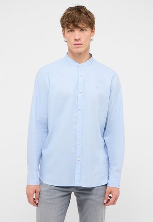 Junger Mann trägt ein hellblaues Hemd mit Mandarin-Kragen und langen Ärmeln sowie hellgraue Jeans, steht vor einem schlichten weißen Hintergrund.