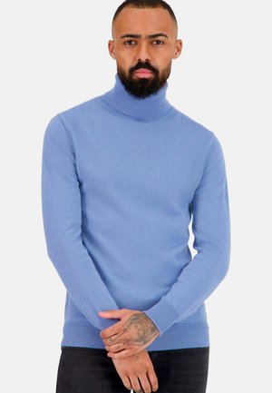 Pull col montant bleu clair, en matériau doux. Design ajusté avec des poignets et un ourlet côtelés, porté avec un jean foncé et les mains croisées.