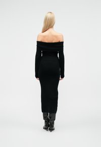 Abito nero lavorato a maglia con scollo a barca, maniche lunghe, silhouette aderente e lunghezza alla caviglia. Indossato con stivali neri al ginocchio.