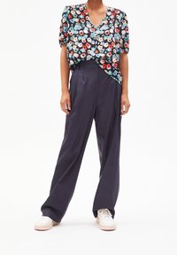 Persona vestida con una blusa de manga corta con estampado floral y escote en V, pantalones de pierna ancha de talle alto en color navy, y zapatillas blancas, de pie frente a un fondo blanco.
