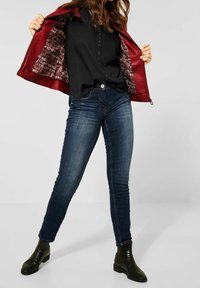 Rote Jacke mit glänzender Oberfläche und gemustertem Innenfutter, getragen über einem schwarzen Oberteil mit dekorativen Knöpfen, kombiniert mit dunkelblauen Skinny-Jeans und schwarzen Ankle-Boots.