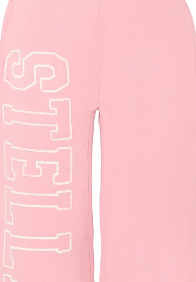 Roze joggingsbroek van zacht materiaal, met grote witte tekst die "STELLA" verticaal langs de linkerkant spelt. Casual ontwerp, losse pasvorm.
