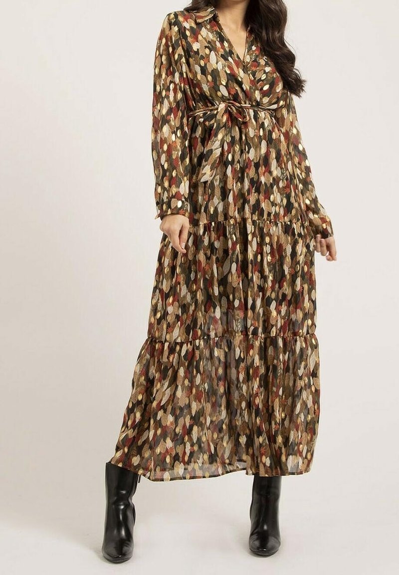 Femme portant une longue robe à motifs aux couleurs des feuilles d'automne, nouée à la taille, associée à des bottines noires, debout devant un fond uni.