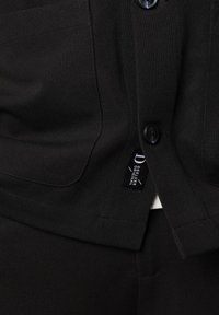 Zwarte textuurgarment met twee knopen, een zak en een label voor "Deluxe Original." De stof lijkt dik en duurzaam.