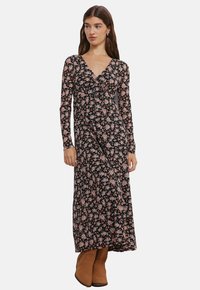 Lang sort blomstret kjole med lange ærmer, prydet med lyserøde og hvide blomster, dyb V-udskæring og en flydende silhuet. Bæres med brune støvler.