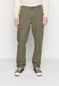 Pantalones cargo de color verde olivo hechos de algodón, con dos grandes bolsillos laterales, un diseño de pierna recta y cierre de botón. Combinados con zapatillas negras.