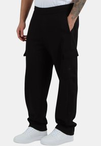 Pantaloni cargo neri con una vestibilità rilassata, dotati di tasche laterali, una texture liscia e una cintura elastica, abbinati a sneakers bianche.