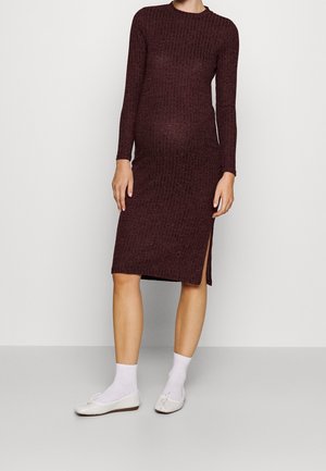 Schwangere Frau in einem knielangen, langärmeligen, bordeauxfarbenen gerippten Kleid mit weißen Socken und weißen Ballerinas, stehend vor einem schlichten Hintergrund.