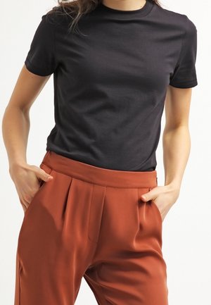 Vrouw draagt een aansluitend zwart T-shirt met korte mouwen, ingestopt in een roestkleurige broek met hoge taille, handen in de zakken.