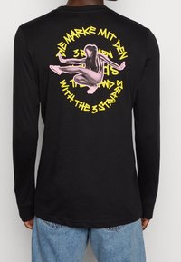 Svart långärmad t-shirt med en stor rosa grafik av en figur och gul text på baksidan. Huvudmaterialet är bomull med en slät textur.