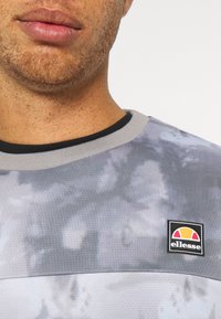 Camisa atlética gris con un patrón de tie-dye, con cuello redondo y el logo de "ellesse" en un parche rectangular en el pecho.