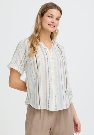 Kvinde med langt bølget hår iført en kortærmet, knappet bluse med lodrette striber og lysebrune bukser, smiler let.