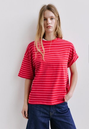 Jonge vrouw met lang blond haar, die een los rood T-shirt met dunne witte horizontale strepen en donkerblauwe spijkerbroek draagt, staand tegen een witte muur.