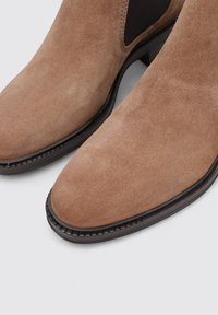 Bottines en suede marron doux avec un bout pointu, des accents élastiques sur les côtés et un talon marron foncé, présentant des coutures détaillées le long de la semelle.