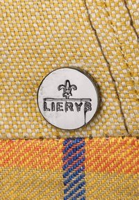 Circular metallic badge with "LIERYS" engraved, featuring a fleur-de-lis symbol, attached to textured yellow and patterned fabric with red and blue accents.