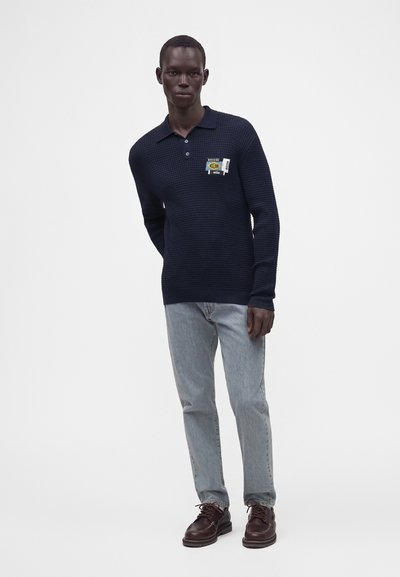 Homme portant un pull en tricot texturé bleu foncé avec une pièce sur la poitrine, jean bleu clair et chaussures à lacets marron foncé, debout sur un fond blanc.